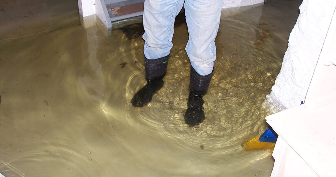A person in jeans and rubber boots stands in a flooded basement, likely needing sewage backup cleanup, with water covering the floor and reflecting the surroundings. Stairs and white walls are visible in the background.