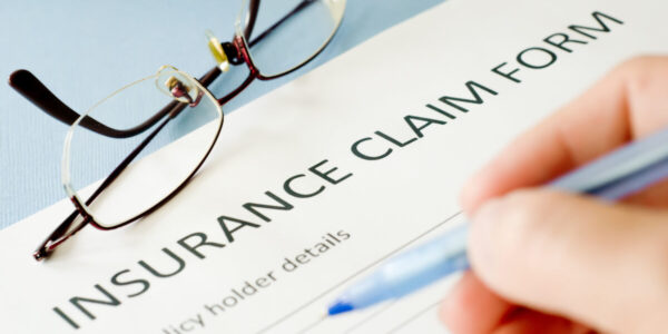 A close-up of a hand filling out an insurance claim form with a pen. Eyeglasses rest on the partially visible form labeled “INSURANCE CLAIM FORM,” highlighting the need for Insurance Assistance Toronto.